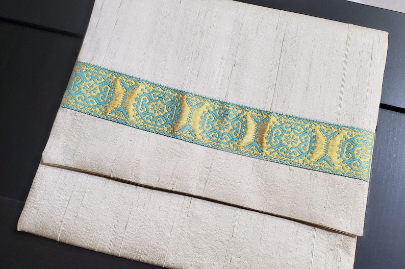 May include: A white fabric envelope with a decorative gold and teal trim. The trim features a repeating geometric pattern.