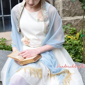 May include: A light blue sheer shawl with floral embroidery and gold trim. The shawl is draped over a white dress, with a matching floral accent. The text "HandmadeByChana" is visible in the image.