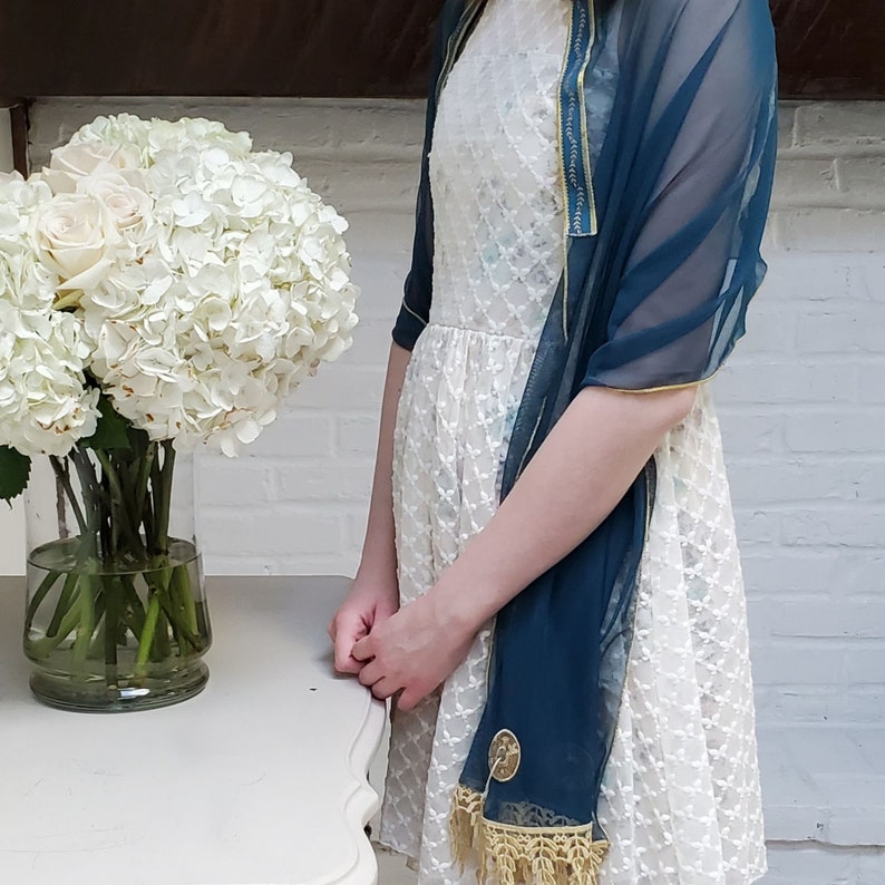 May include: A white dress with a blue and gold embroidered shawl. The dress has a lace pattern and the shawl has a fringe trim.