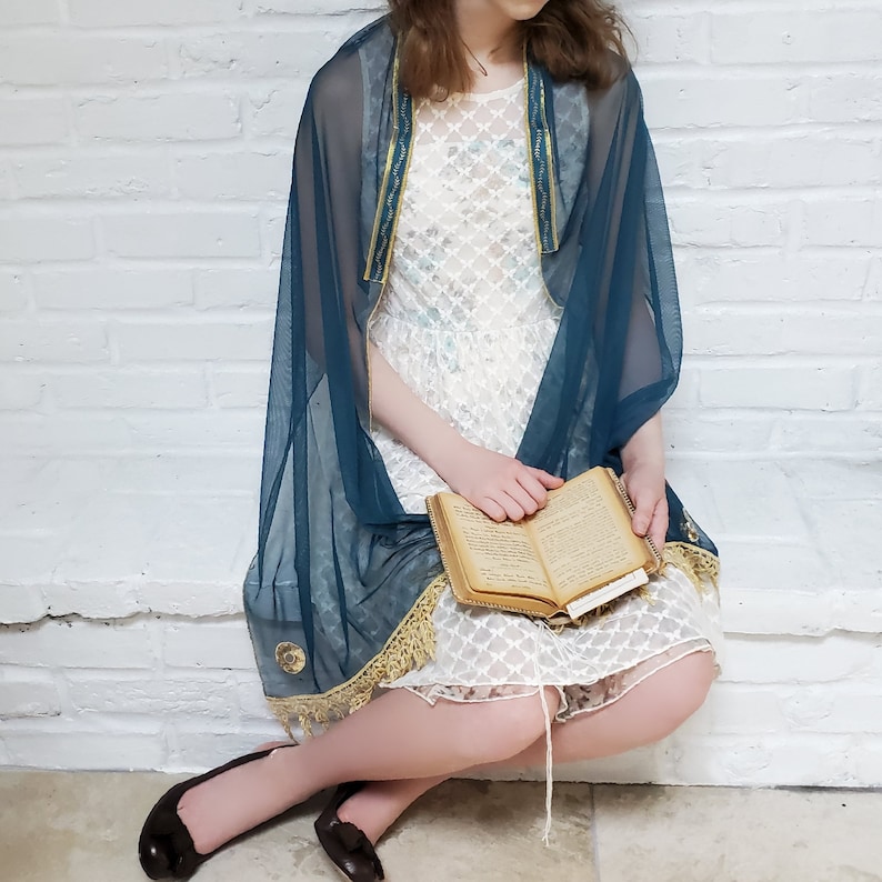 May include: A woman wearing a teal blue sheer shawl with gold trim over a white lace dress. She is holding a book with a gold cover.