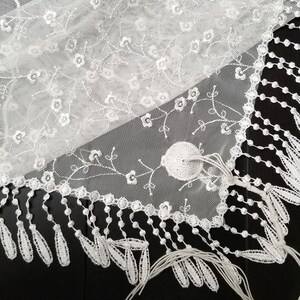 May include: Close-up of a white lace fabric with floral embroidery and a scalloped edge. The delicate lace features small flower and vine details, with a fringe of teardrop-shaped tassels along the border. The fabric is on a black surface.