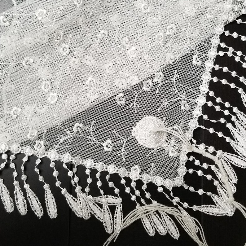 May include: Close-up of a white lace fabric with floral embroidery and a scalloped edge. The delicate lace features small flower and vine details, with a fringe of teardrop-shaped tassels along the border. The fabric is on a black surface.