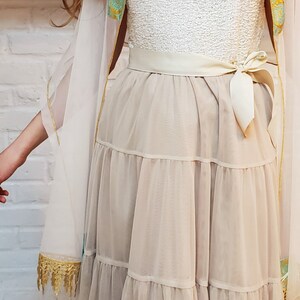 May include: A white dress with a tiered skirt and a white sash tied in a bow at the waist. The dress is layered over a sheer white fabric with gold trim.