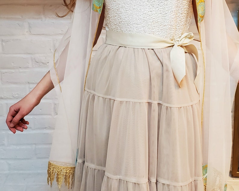 May include: A white dress with a tiered skirt and a white sash tied in a bow at the waist. The dress is layered over a sheer white fabric with gold trim.