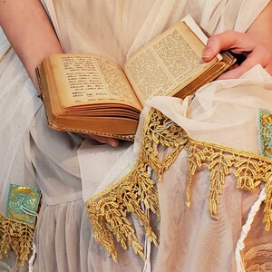 May include: A white shawl with gold and turquoise trim. The shawl is decorated with intricate lace and has a book resting on top.