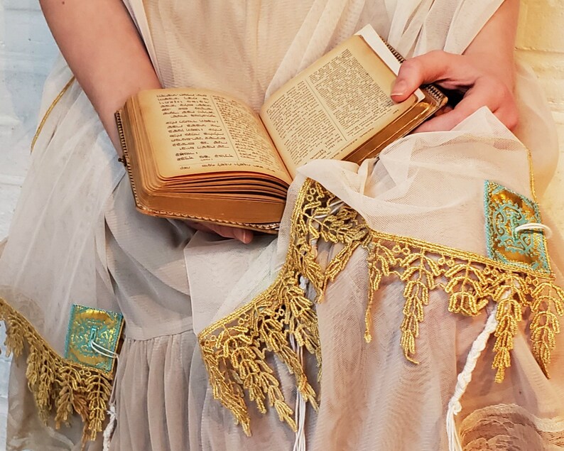 May include: A white shawl with gold and turquoise trim. The shawl is decorated with intricate lace and has a book resting on top.
