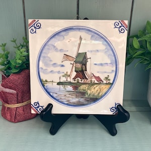 May include: Square ceramic tile depicting a Dutch windmill scene within a blue circular border. The windmill has green and brown accents, with a water and landscape background. Decorative corner designs in red and blue.