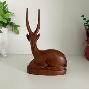 May include: A wooden sculpture of a resting antelope with long, upright horns. The antelope is carved from a rich, dark brown wood and sits on a rounded base. The sculpture is a decorative item, suitable for home decor.