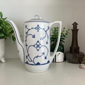 May include: A white porcelain coffee pot featuring a tall cylindrical body, a curved handle, and a long spout. The exterior is decorated with a delicate blue filigree pattern of interconnected vines and geometric floral motifs.