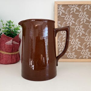May include: A shiny, brown ceramic jug with a handle and a pouring spout. The jug is on a white surface, with a framed lace artwork and potted plants behind it. A classic kitchenware item.