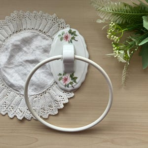 May include: A white ceramic towel ring featuring a scalloped oval wall mount decorated with pink rose floral patterns and green leaves. The circular ring is attached to the centre of the mount, providing a functional bathroom fixture.