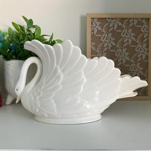 May include: A white ceramic swan planter with a detailed wing and feather design. The swan has a curved neck and a small, pointed beak. The planter is on a white surface, with greenery and a framed lace artwork in the background.