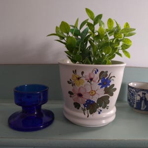 May include: A white ceramic planter with a floral design featuring pink, yellow, blue, and green flowers. The planter is filled with a green leafy plant and is sitting on a light blue surface.