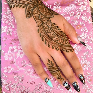 May include: A hand with intricate henna designs on the wrist and fingers. The henna is a dark brown color and the design is floral and geometric. The hand is resting on a pink fabric with a floral pattern.