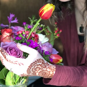 May include: A hand with intricate henna designs holds a bouquet of flowers, including a purple rose, a yellow and red tulip, and other purple and pink flowers. The hand is wearing a ring. The flowers are in a clear glass vase. The background is a blurry image of a person with long dark hair.