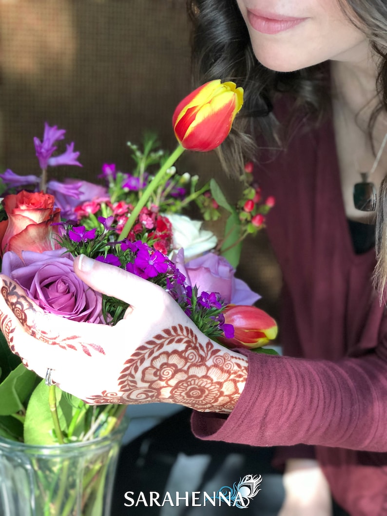 May include: A hand with intricate henna designs holds a bouquet of flowers, including a purple rose, a yellow and red tulip, and other purple and pink flowers. The hand is wearing a ring. The flowers are in a clear glass vase. The background is a blurry image of a person with long dark hair.