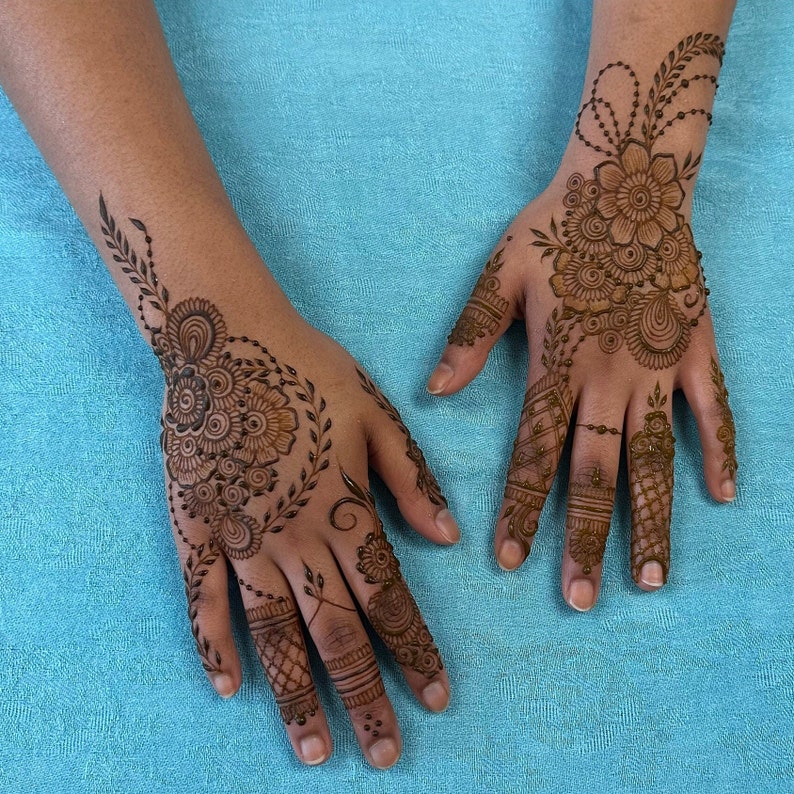 May include: Close-up of two hands with intricate henna designs. The designs feature floral patterns, swirls, and dots. The henna is a dark brown color and is applied to the back of the hands and fingers.
