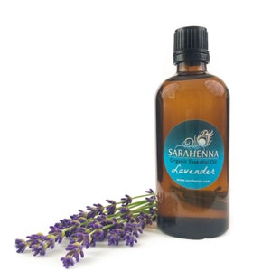 May include: A brown glass bottle of organic lavender essential oil with a black cap. The bottle has a blue and white label with the text "SARA HENNA Organic Essential Oil Lavender www.sarahenna.com". A bunch of lavender flowers are in the foreground.