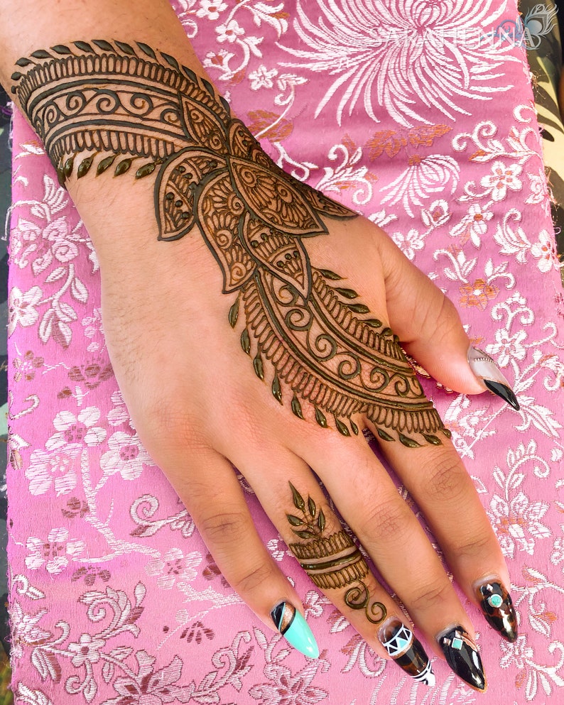 May include: A hand with intricate henna designs on the wrist and fingers. The henna is a dark brown color and the design is floral and geometric. The hand is resting on a pink fabric with a floral pattern.