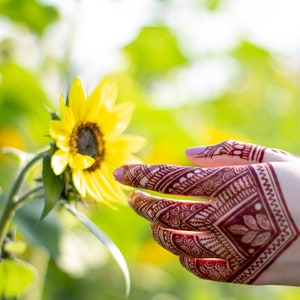 May include: A hand with intricate henna designs reaches towards a bright yellow sunflower in a field of green.