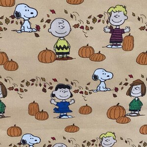 May include: Tan fabric featuring a repeating pattern of cartoon characters, pumpkins, and autumn leaves. The characters include Snoopy, Charlie Brown, Lucy, and others. The pumpkins are orange, and the leaves are red, orange, and brown.