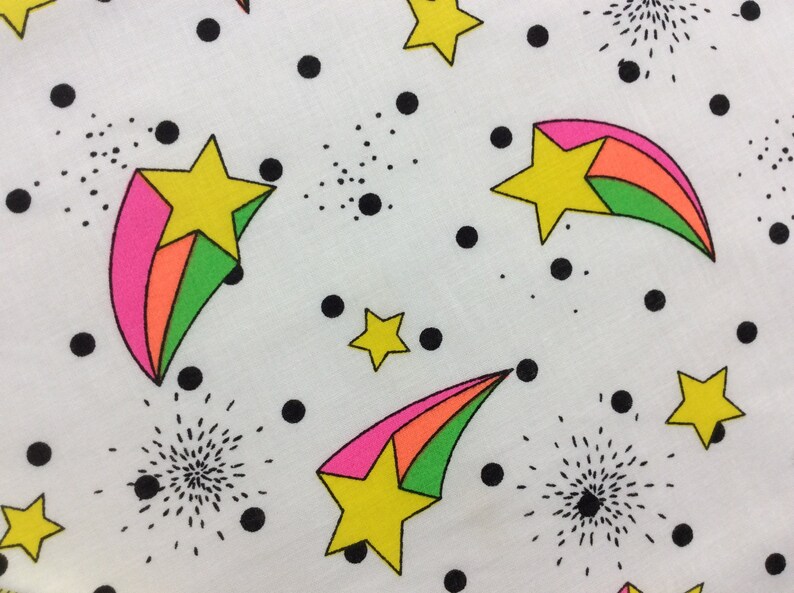 May include: White fabric with a repeating pattern of black dots, green, pink, and orange shooting stars with yellow stars in the center.