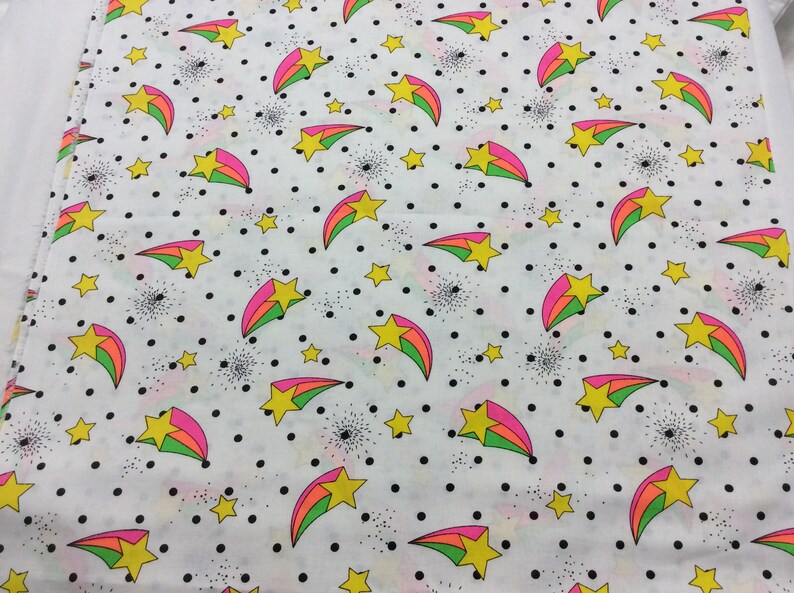 May include: White fabric with a repeating pattern of yellow stars and pink, green, and blue shooting stars on a white background with black polka dots.