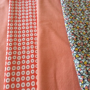 May include: Three fabric swatches in shades of coral. The left swatch has a white polka dot pattern, the middle swatch is solid coral, and the right swatch has a geometric pattern of overlapping circles in shades of coral, yellow, and black.