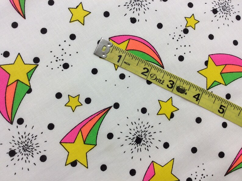 May include: White fabric with a repeating pattern of black dots, green, pink, and yellow shooting stars, and black firework designs. A yellow measuring tape with black markings reads "1 inch, 2 Dritz, 3, 4, 5".