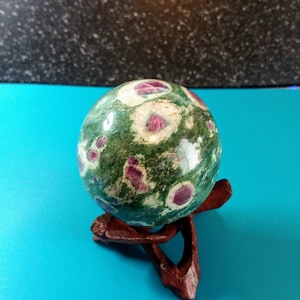 May include: Green and pink ruby zoisite sphere on a wooden stand. The sphere is polished and has a variety of colors and patterns.