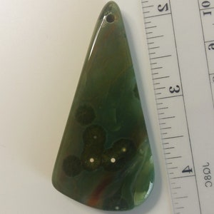 May include: A green, brown, and white agate pendant with a hole drilled at the top. The pendant is shaped like a triangle and has a smooth, polished surface.
