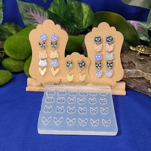 May include: A set of 24 cat face silicone molds for making resin earrings. The molds are clear and show the cat face design. There are two wooden display stands with cat face earrings in various colors.