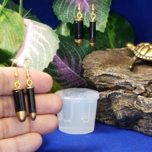 Pode incluir: Um par de brincos de ouro em forma de bala com detalhes pretos. Os brincos são exibidos em uma mão e em uma superfície de madeira com uma pequena figura de tartaruga.