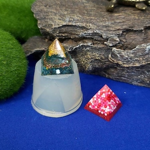May include: Two resin pyramid molds, one clear and one filled with red and white glitter resin. The clear mold is sitting on a wooden surface.