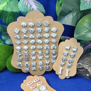 May include: A set of 36 dog-themed stud earrings displayed on a brown card with the text "Doms Creations". The earrings feature various dog breeds, including a pug, a bulldog, a chihuahua, and a dachshund. There are also a few earrings with paw prints and bones.