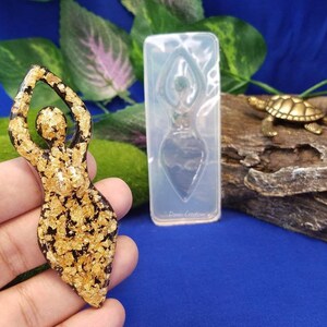 以下が含まれることがあります： 青い背景に、腕を上げた女性の金色のレジン製フィギュア。フィギュアは金箔でできており、手のひらに乗っています。画像には、同じフィギュアの透明なシリコンモールドも写っています。