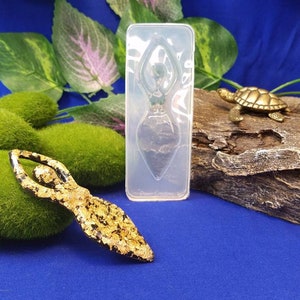 以下が含まれることがあります： 樹脂工芸品を作るための透明なシリコンモールド。モールドは、腕を頭上に上げたヨガポーズをとる女性の形をしています。モールドは、緑の植物と小さな金の亀の置物のある青い表面の上にあります。