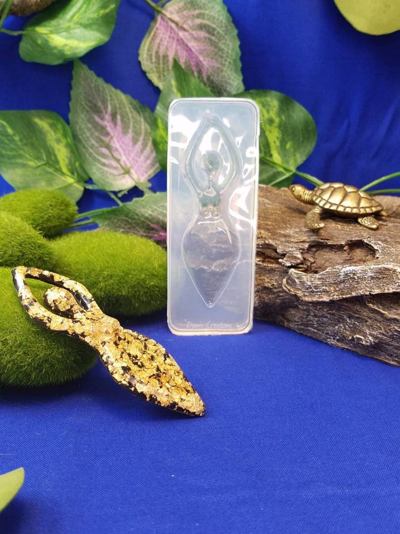 以下が含まれることがあります： 樹脂工芸品を作るための透明なシリコンモールド。モールドは、腕を頭上に上げたヨガポーズをとる女性の形をしています。モールドは、緑の植物と小さな金の亀の置物のある青い表面の上にあります。