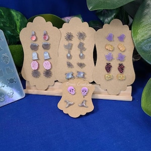 May include: A set of Halloween-themed earrings displayed on a wooden stand. The earrings are made of resin and feature various designs, including skulls, witches' hats, ghosts, spiders, and cauldrons. The earrings are all in shades of purple, black, and gold.