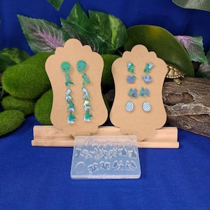 Pode incluir: Um conjunto de dois expositores de brincos com vários brincos de resina verde e azul. Os brincos são exibidos em cartões de papelão marrom. Também está incluído um molde de silicone transparente com várias formas.