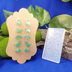 May include: A set of green dinosaur earrings and a clear silicone mold with dinosaur shapes. The earrings are on a brown card with a scalloped edge. The mold has the text "Dino Creations" on the bottom.