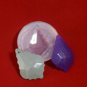 May include: Two clear resin pendants in the shape of a teardrop with a faceted design. One pendant is light green and the other is purple. The pendants are sitting on a red surface next to a silicone mold.