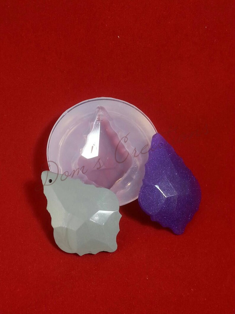 May include: Two clear resin pendants in the shape of a teardrop with a faceted design. One pendant is light green and the other is purple. The pendants are sitting on a red surface next to a silicone mold.