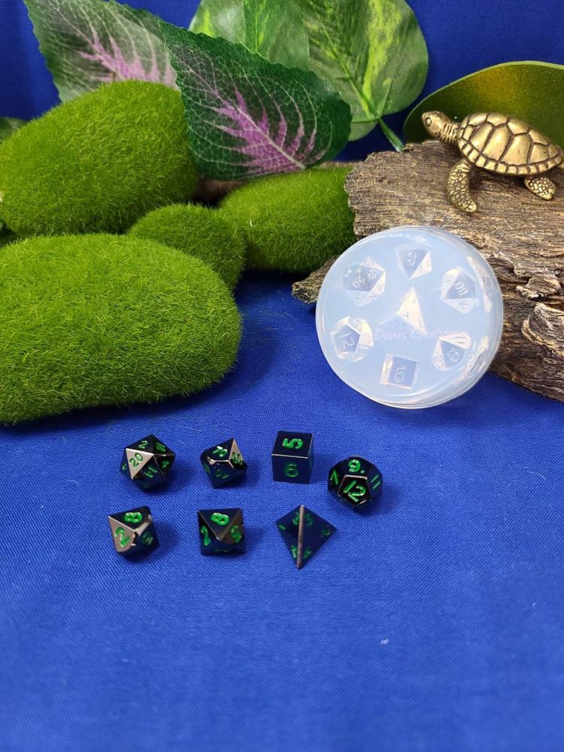 May include: A set of seven black metal dice with green numbers. The dice are arranged in a circle on a blue surface. A clear plastic mold for dice is also visible in the image.