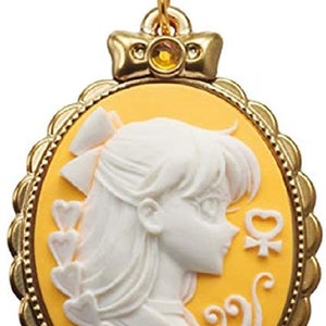 May include: A gold-tone charm featuring a cameo of a white silhouette of a woman with a yellow background. The charm is in the shape of an oval with a decorative border. The charm has a small gold-tone loop at the top for attaching to a chain or bracelet.