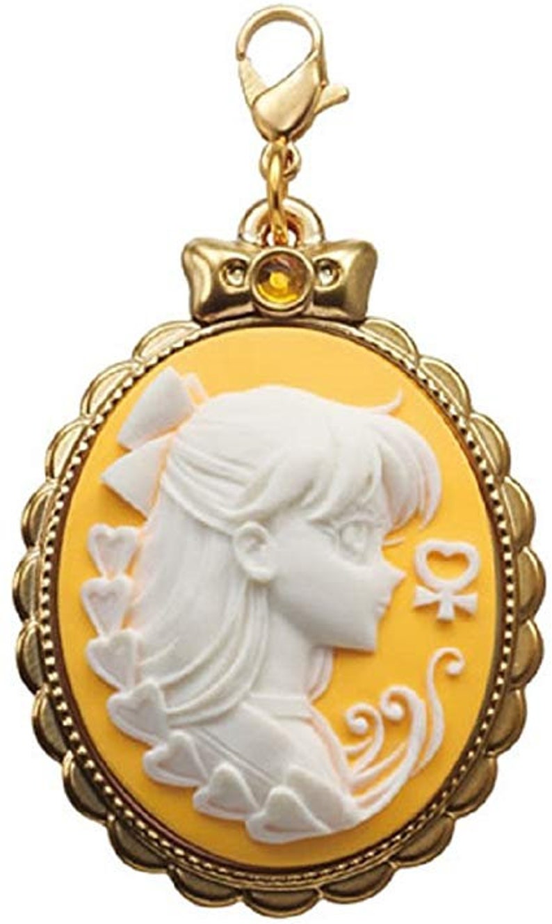 May include: A gold-tone charm featuring a cameo of a white silhouette of a woman with a yellow background. The charm is in the shape of an oval with a decorative border. The charm has a small gold-tone loop at the top for attaching to a chain or bracelet.
