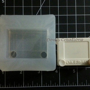 May include: A white plastic mold for making a toy Etch A Sketch. The mold is 3 inches wide and 4 inches long. The mold has a rectangular opening for the drawing area and two round openings for the knobs.