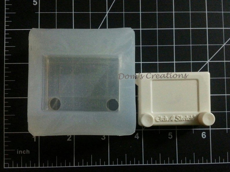 May include: A white plastic mold for making a toy Etch A Sketch. The mold is 3 inches wide and 4 inches long. The mold has a rectangular opening for the drawing area and two round openings for the knobs.