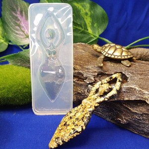 以下が含まれることがあります： 樹脂工芸品を作るための透明なシリコンモールド。モールドは、頭上に腕を上げた女性のフィギュアの形をしています。モールドは、同じフィギュアの金色の樹脂製の鋳造物の隣に示されています。