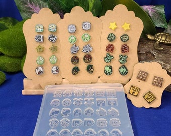 Può includere: Un set di 36 stampi per orecchini a tema Nintendo, con personaggi e oggetti della serie Super Mario Bros. Gli stampi sono in silicone trasparente e sono esposti su un pannello di legno con orecchini di esempio.